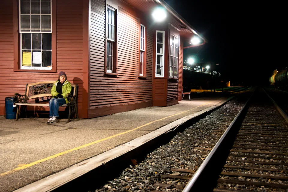 Womanin yellow jacket sitting on bench at outdoor train station with red wood depot building at night.